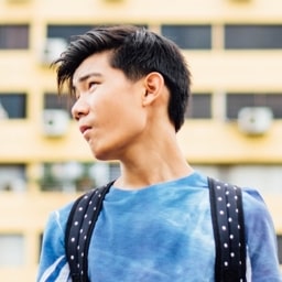 Profile shot of Aden Allan, wearing a bright blue top while carrying a dark blue and white spotted rucksack. The shot was taken with a blurred backdrop of a block of council flats.