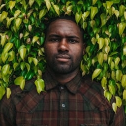 Profile shot of Drake Heaton, wearing a dark brown and red shirt, taken with his front body embedded inside the front of a hedge.